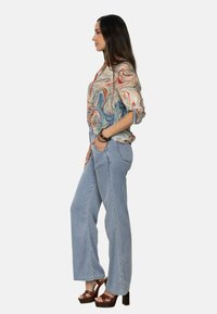 Blusa de mármol multicolor con mangas largas, vaqueros acampanados de color azul claro y tacones de plataforma marrones, vista de lado contra un fondo blanco.