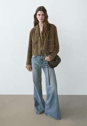 Giovane donna con capelli lunghi che indossa una giacca corta in camoscio marrone, top a stampa leopardata, jeans blu a gamba larga e porta una borsa nera a tracolla.