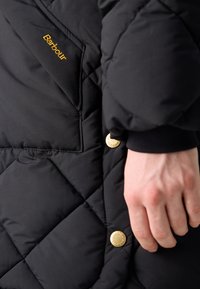 Zwarte gestikte jack met gouden hardware en geborduurd "Barbour"-logo. Heeft een gladde textuur en een getailleerd silhouet met een ronde manchet.