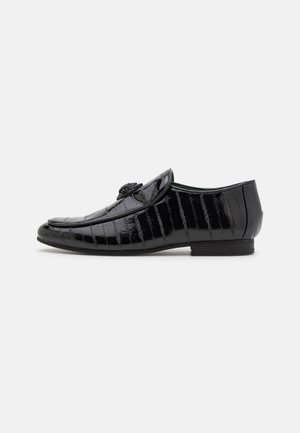 Mocassin en cuir verni noir avec texture crocodile, doté d'une sangle décorative et d'un talon bas, présenté en profil latéral sur fond blanc.