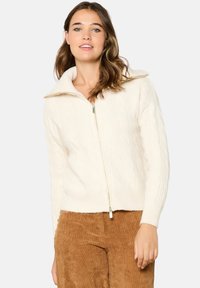 Kremo spalvos megztinis su kabeliniu raštu, aukštu apykakle ir užtrauktuku, derinamas su rudais kilpiniais kelnėmis. Minkšta tekstūra ir pritaikytas dizainas.