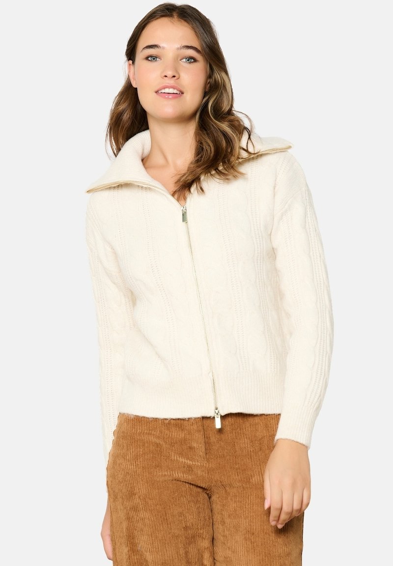 Kremo spalvos megztinis su kabeliniu raštu, aukštu apykakle ir užtrauktuku, derinamas su rudais kilpiniais kelnėmis. Minkšta tekstūra ir pritaikytas dizainas.