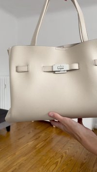 Beige skinnveske med doble håndtak, en sølvklips og en teksturert overflate. Har minimalistisk design og strukturert form.