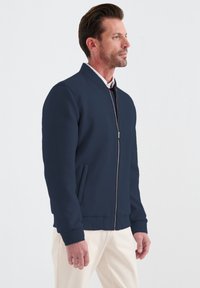 Marinblå bomberjacka med dragkedja i fronten, ribbade manschetter och nederkant samt sidofickor. Slät tyg med en ren, minimalistisk design.