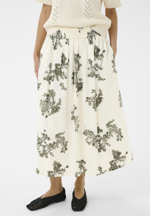 Frau trägt einen cremefarbenen Midirock mit schwarzen Blumenmustern, kombiniert mit schwarzen gewebten Slippern und einem cremefarbenen Strickpullover.