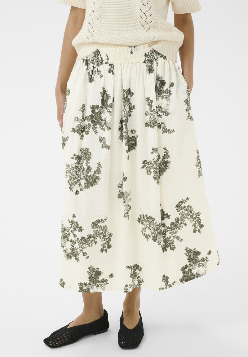 Donna che indossa una gonna midi color crema con motivi floreali neri, abbinata a scarpe slip-on nere intrecciate e a un maglione lavorato a maglia color crema.