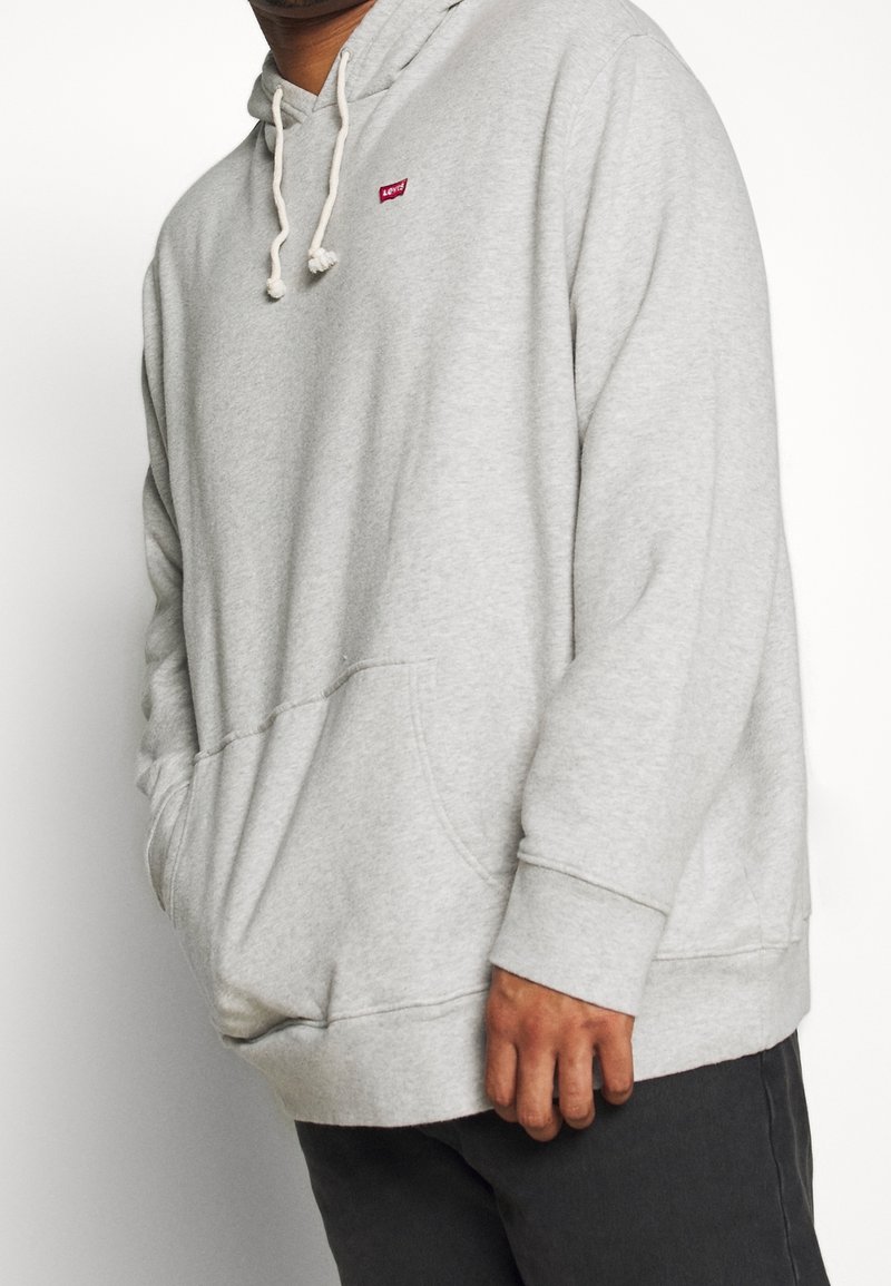 Personne portant un sweat à capuche gris clair avec cordons et un petit logo rouge Levi's sur la poitrine, associé à un pantalon foncé.