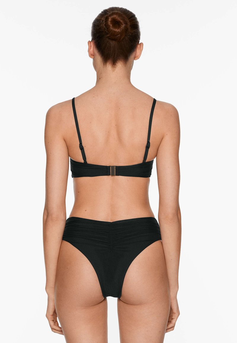 Mujer con el cabello recogido en un moño que lleva un traje de baño de dos piezas negro, vista desde atrás sobre un fondo blanco liso.