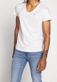 Man klädd i en enfärgad vit V-ringad t-shirt med en liten logotyp och ljusblå jeans, som visar en tatuering på sin högra underarm.