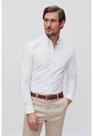 Hvid skjorte med knapper og lange ærmer, parret med lyse beige bukser. En brun fletbælte tilføjer tekstur. Neutral baggrund.