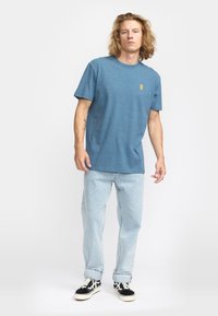 Camiseta de algodón azul con cuello redondo y mangas cortas, que presenta un pequeño gráfico amarillo en el pecho. Combinada con jeans azules claros.