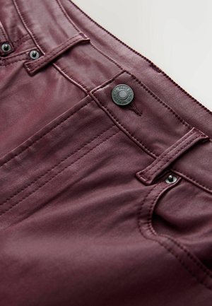 Burgundy jeans med en slät yta, femfickorsdesign, svart metallknapp och kontrasterande sömdetaljer nära fickorna.