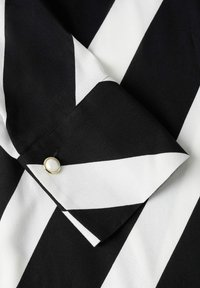 Blouse à rayures noires et blanches avec un col plié et un grand bouton rond en perle sur le poignet, confectionnée dans un tissu lisse et léger.