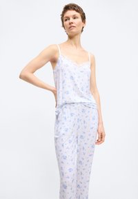 Γυναίκα που φορά άσπρο τοπ και παντελόνι με μικρό μπλε ανθισμένο σχέδιο, όρθια με το ένα χέρι στη μέση της μπροστά σε απλό φόντο.