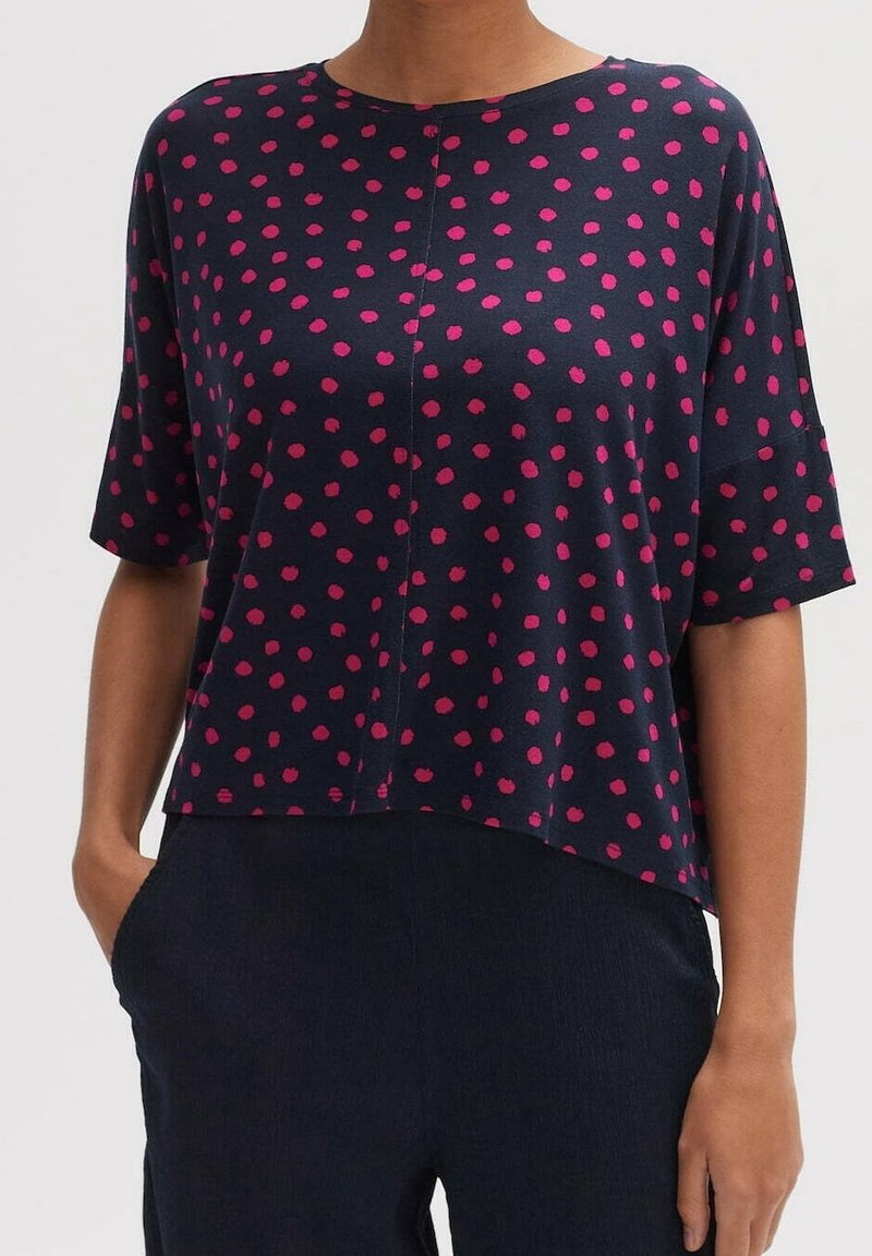 Femme portant une chemise bleu marine à manches courtes avec des pois roses et un pantalon noir, debout avec une main dans une poche.