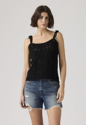 Mujer con cabello corto y castaño, vestida con una parte superior negra sin mangas tejida a crochet y pantalones cortos vaqueros azules desgastados sobre un fondo liso.