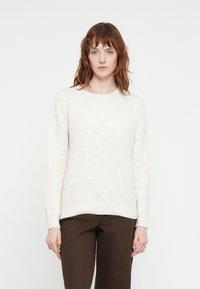 Cream knitted sweater with a textured pattern, featuring a round neckline and long sleeves, paired with brown trousers.