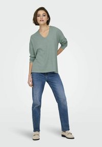 Lichtgroene, lange mouw V-hals top, gecombineerd met blauwe spijkerbroek en beige schoenen, wat zorgt voor een casual outfit. Zachte stof en een comfortabele pasvorm.