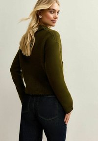 Oliwkowy sweter dziany o cropped fasonie i prążkowanym wykończeniu, zestawiony z ciemnymi dżinsami. Gładka faktura i luźny krój.