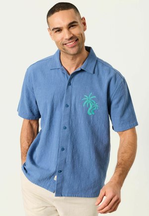 Mann lächelt, trägt ein blaues kurzärmliges Hemd mit grünem Palmenmuster, beige Hose, steht vor einem schlichten Hintergrund.