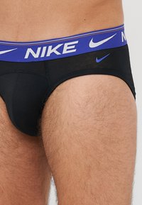 Schwarze Nike-Unterwäsche mit einem blauen Taillenbund, der den weißen "NIKE"-Text und das Logo zeigt. Weicher Stoff, enganliegendes Design und elastischer Taillenbund für zusätzlichen Komfort.