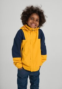 Veste coupe-vent jaune et marine avec capuche, poches zippées et poignets élastiques, portée sur un jean en denim. Enfant souriant, les mains dans les poches.
