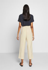 Kvinna med långt mörkt hår som bär en marinblå topp, beige capribyxor och svarta slip-on skor, står vänd mot en enkel ljusgrå bakgrund.