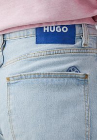 Vaqueros de mezclilla azul claro con un parche de cuero azul etiquetado como "HUGO" en la cintura, con costuras amarillas y un pequeño logo azul en el bolsillo trasero.