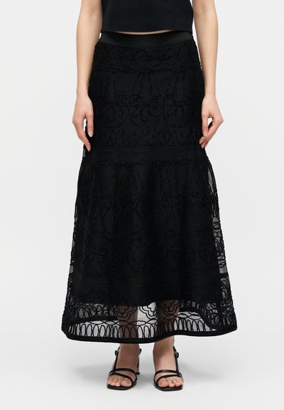 Femme portant une jupe midi en dentelle noire avec des motifs floraux et des sections transparentes, associée à des sandales à talons noires à brides.