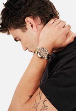 Ung mand med mørkt hår holder om nakken med venstre hånd og viser et guld- og sølvfarvet armbåndsur, synlig arm-tatovering og sort skjorte.