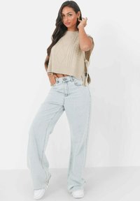 Crop top en tricot beige avec des motifs texturés, associé à un jean large bleu clair et des baskets blanches. Le mannequin se tient avec une main sur la hanche.