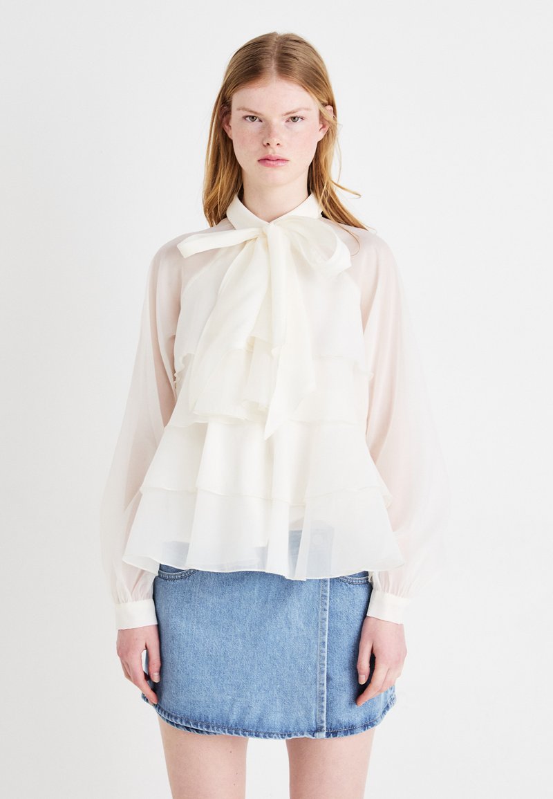 Hvit, transparent bluse med lagdelte volanger og krage med sløyfe, kombinert med et lys blått denim mini-skjørt. Lange, svulmende ermer.