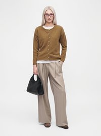 Femme aux longs cheveux blonds, portant des lunettes, un cardigan marron, un pantalon large beige, des chaussures marron, tenant un sac à main noir, debout sur un fond blanc.