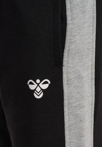 Svarta och grå sweatpants med en mjuk textur, sidostrepo-design och en vit grafisk logotyp på vänster ben.