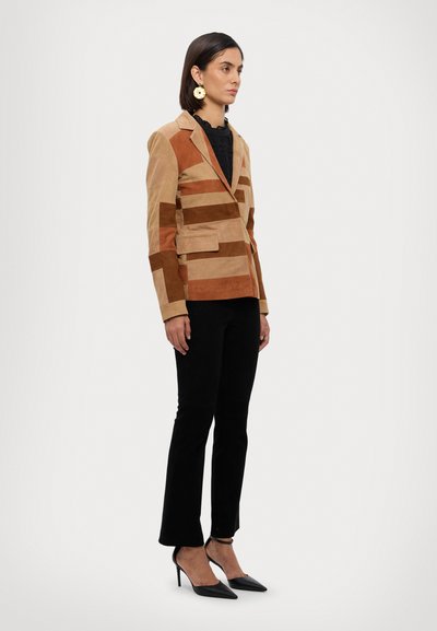 Blazer in suede con un motivo geometrico in tonalità beige e marrone, dotato di chiusura a un bottone e due tasche frontali. Abbinato a pantaloni neri.