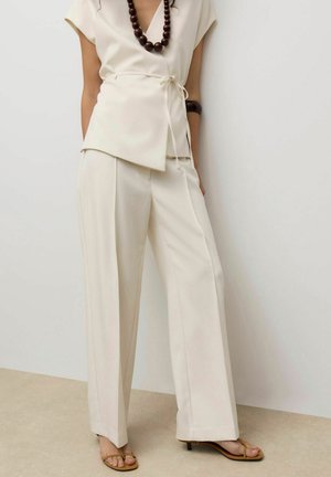 Femme portant un haut cache-cœur crème et un pantalon large taille haute, accessoirisée d'un collier et d'un bracelet en bois foncé, debout contre un mur blanc.