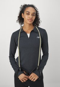 Μακρυμάνικη αθλητική μπλούζα σε σκούρο γκρι χρώμα με μισό φερμουάρ, διακοσμημένη με το σήμα της New Balance στο στήθος και τρύπες για τον αντίχειρα στα μανίκια.