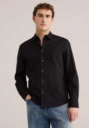 Junger Mann mit lockigem Haar, der ein schwarzes Hemd mit Knöpfen und blaue Jeans trägt, mit einer Hand in der Tasche vor einem einfarbigen Hintergrund steht.