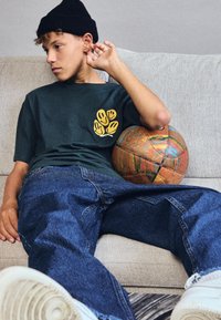Adolescente con gorro negro, camiseta oscura con gráfico amarillo, jeans azul holgados, sentado en un sofá sosteniendo un balón de baloncesto desgastado, mirando hacia un lado.