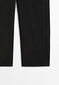 Schwarze Denim-Jeansbeine, von der Mitte des Oberschenkels abwärts, gerade geschnitten, vor weißem Hintergrund.