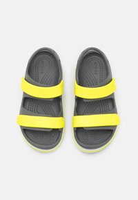 Harmaat sandaalit, joissa on kaksi keltaista hihnaa, ja joissa on teksturoitu pohja sekä logo. Kevyt muotoilu paksulla reunalla mukavuutta varten.