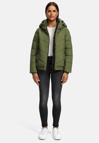 Olijfgroene gewatteerde jas met een capuchon, voorzien van een ritssluiting aan de voorkant, elastische manchetten en een subtiel stiksels patroon. Gedragen over een witte top en zwarte jeans.