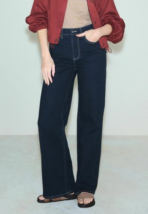 Osoba ubrana w granatowe jeansy z szerokimi nogawkami, beżową bluzkę, czerwoną kurtkę i czarne sandały, stojąca na jasnej podłodze z jedną ręką w kieszeni.