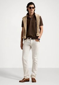 Brun pikétröja under en beige kabelstickad tröja, vita jeans, bruna skor och solglasögon. Har en klocka på vänster handled.