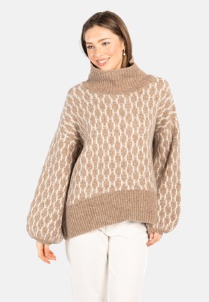 Smilende kvinne iført beige mønstret høyhalset genser med ballongermer og hvite bukser mot ensfarget bakgrunn.