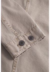 Material din bumbac bej cu un manșon pliat, decorat cu două nasturi gri închis. Detalii ale cusăturilor vizibile și o textură netedă.