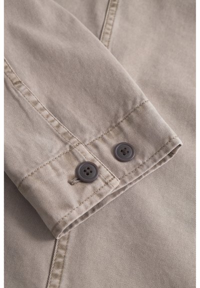 Beige katoenen stof met een omgevouwen manchet waarop twee donkergrijze knopen zijn bevestigd. Zichtbare stikseldetails en een gladde textuur.