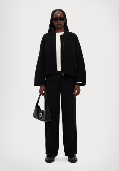 Black cropped jacket with a zipper, button pockets, worn over a cream top and paired with black trousers, black sunglasses, and a handbag.