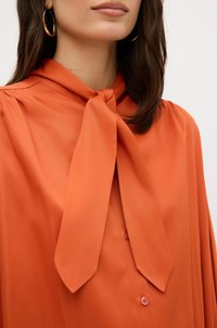Orange Bluse aus glattem Stoff mit einer Krawatte am Hals, Knopfdetails und dezenten vertikalen Streifen für Textur.