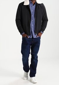 Svart bomberjacka med vit fårsullskrage, blårutigt skjorta under, mörkblå jeans och vita sneakers.